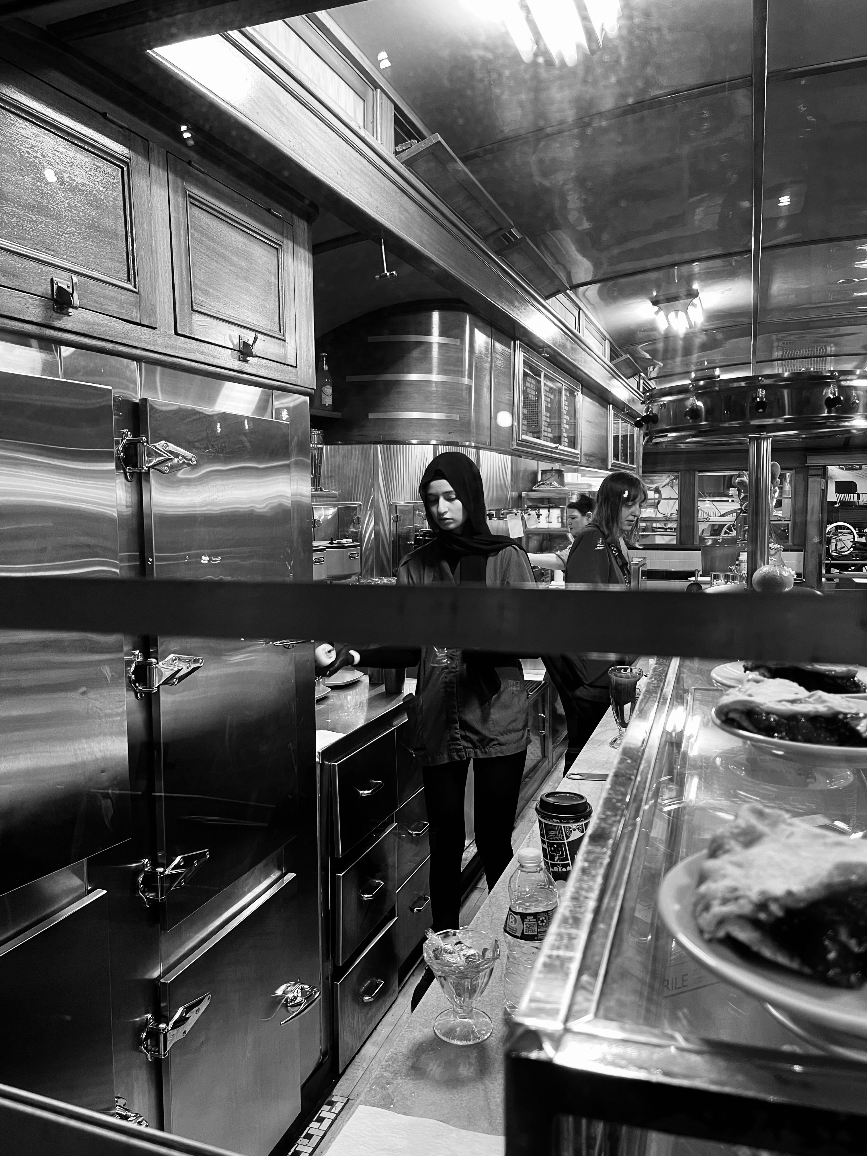 Behind the Scenes — view through a diner counter, chrome and stainless steel surfaces, young woman in center frame, classic American diner, street documentary photography by Craig Moyers