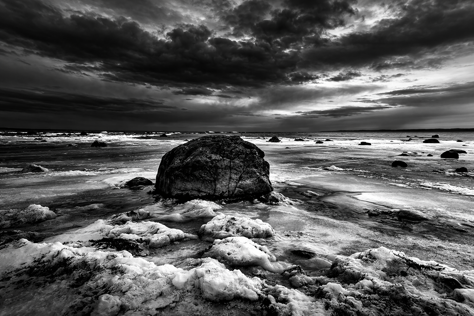 Boulder on frozen shore — ice, snow, and storm clouds over water, fine art black and white landscape by Craig Moyers