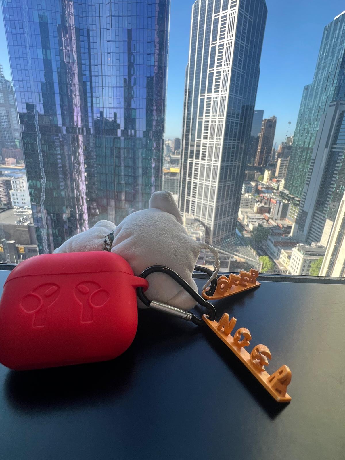 Dual Name Keychain showing OFER and NOSGA, clipped to an AirPods case with a city skyline in the background