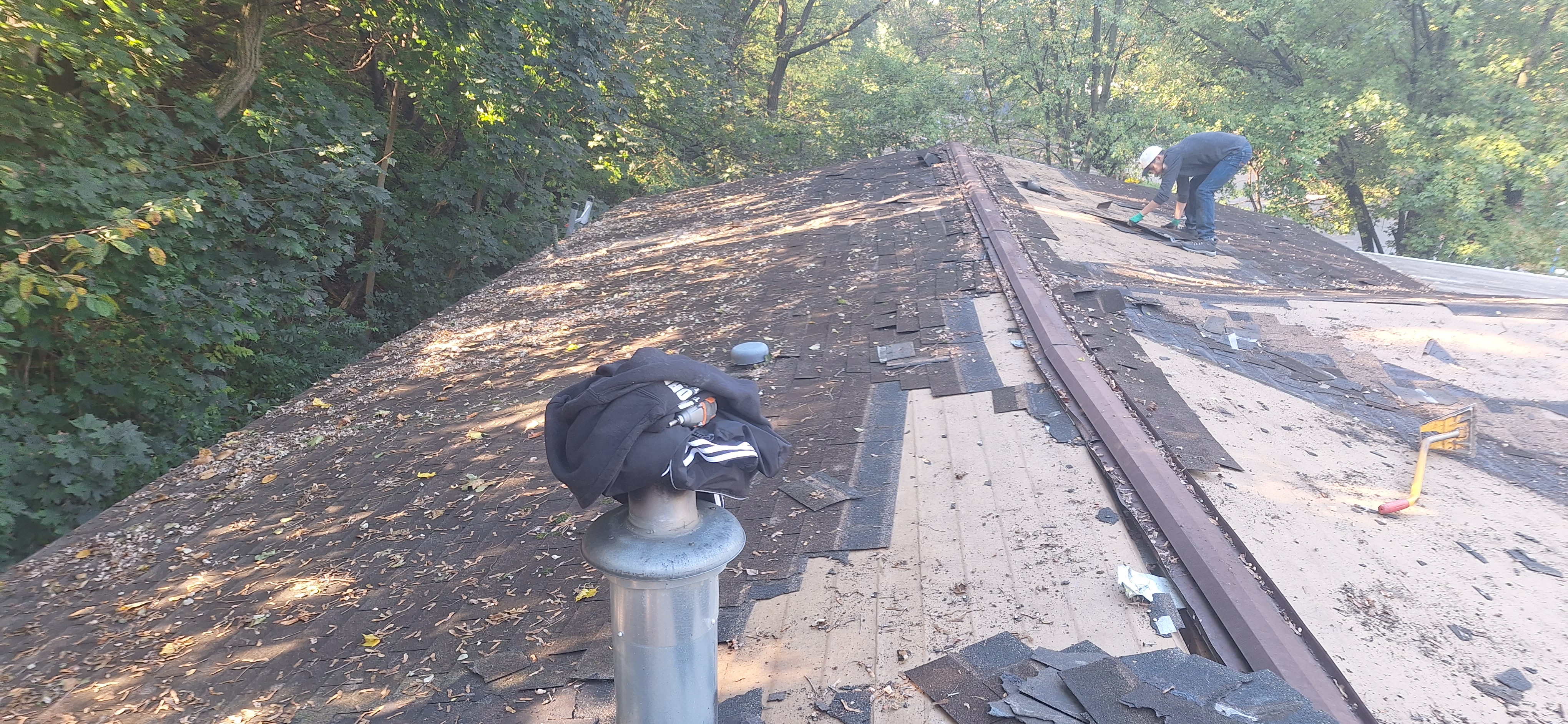 Crew member stripping shingles on large roof tear-off in Pennsylvania — ridge vent removed, old felt visible, vent pipe in foreground