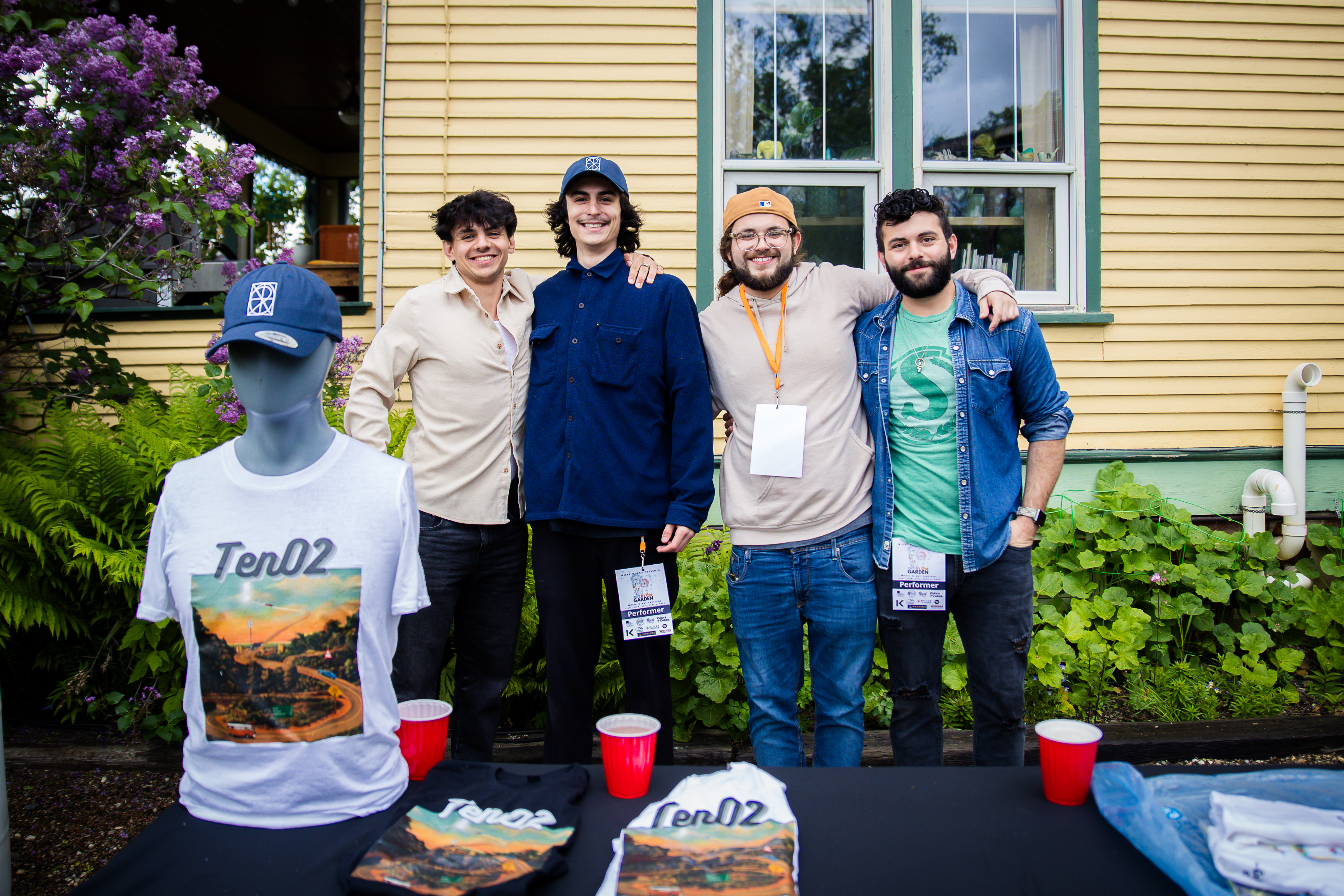 Ten02 band with merch table — four performers with lanyards, farmhouse in the background
