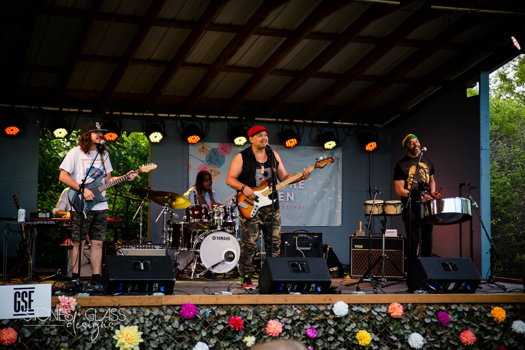 Earl Perrera and The Steadies performing on the outdoor stage at Art in the Garden — flowers lined along the front of the stage