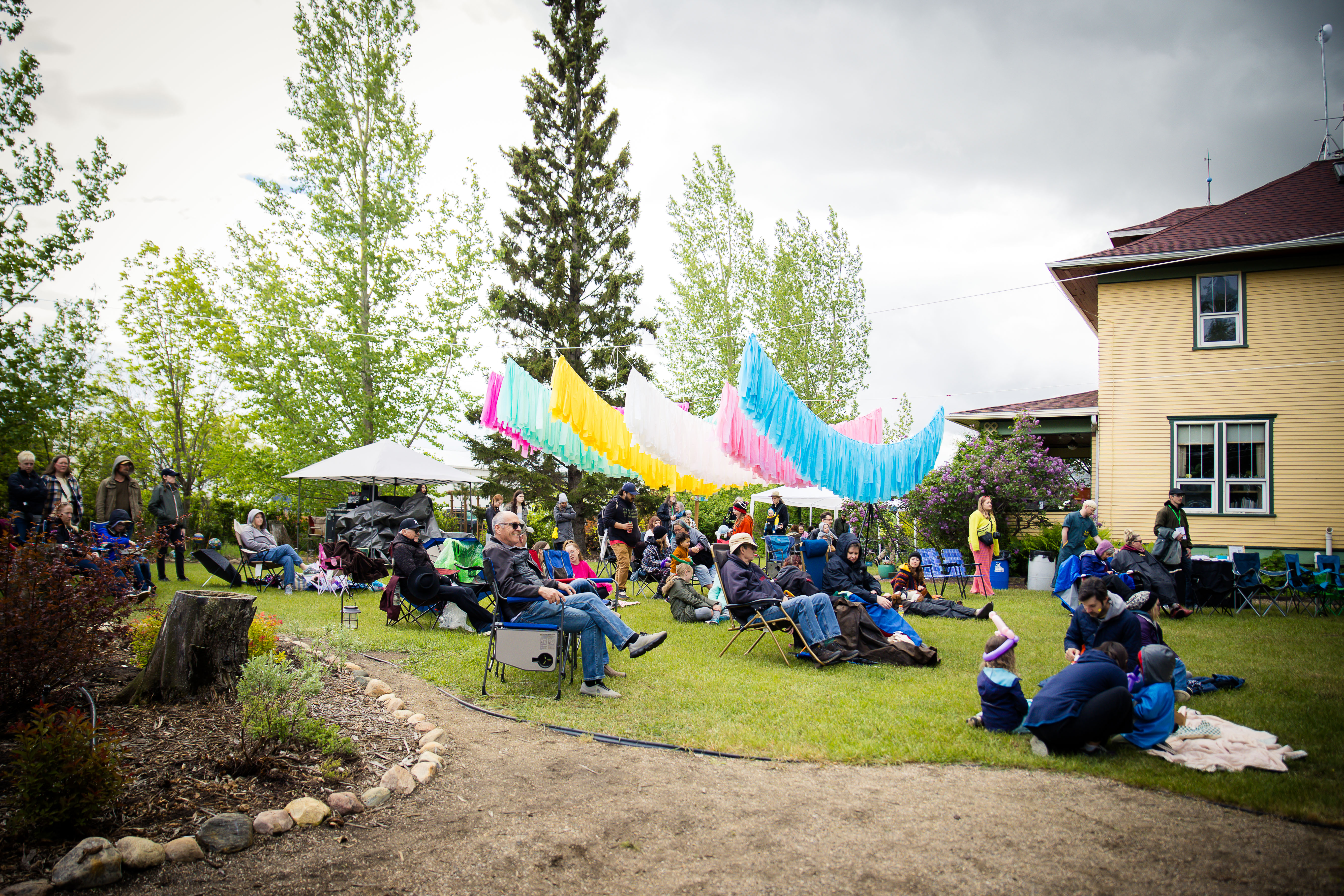 Wide shot of festival crowd on the lawn at Art in the Garden 2026 — chairs, blankets, streamers overhead