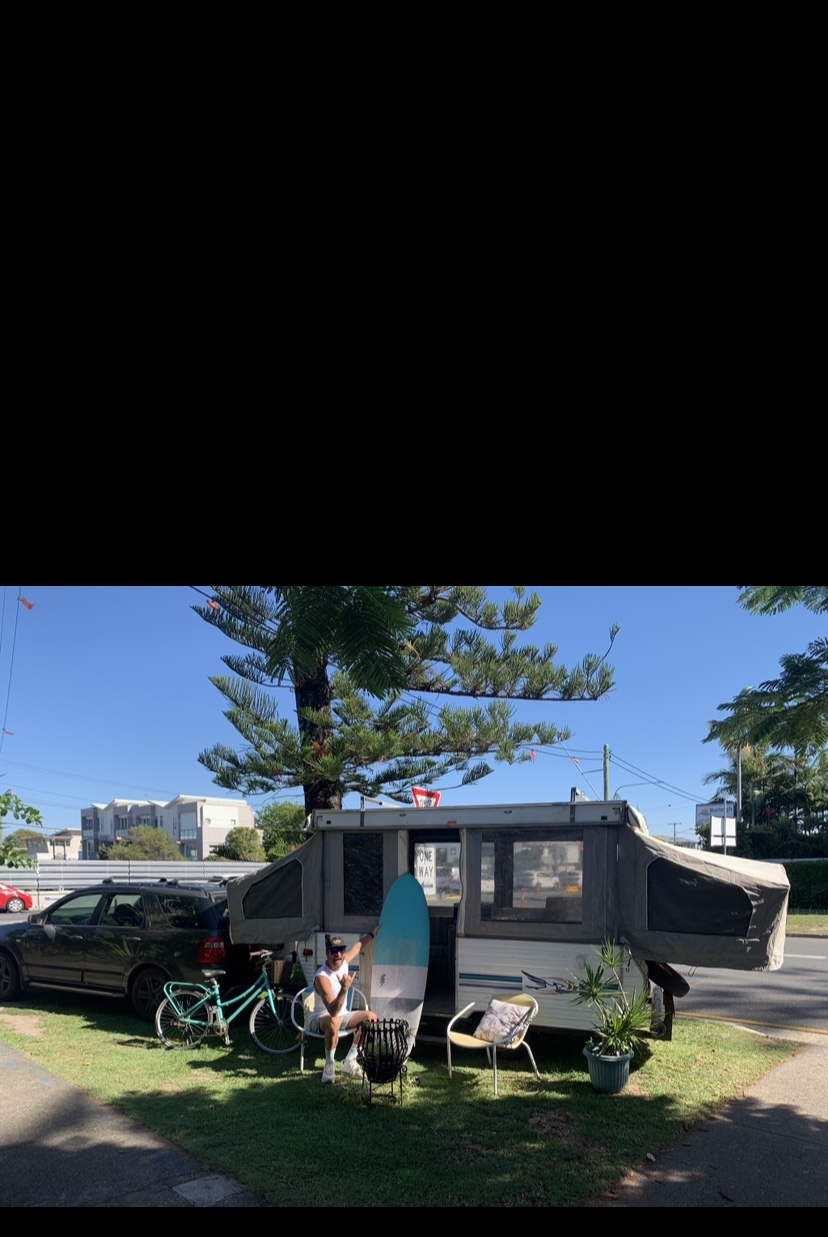 Konstantin outside a pop-up caravan with surfboard and bike — graduated from tent to caravan, still building