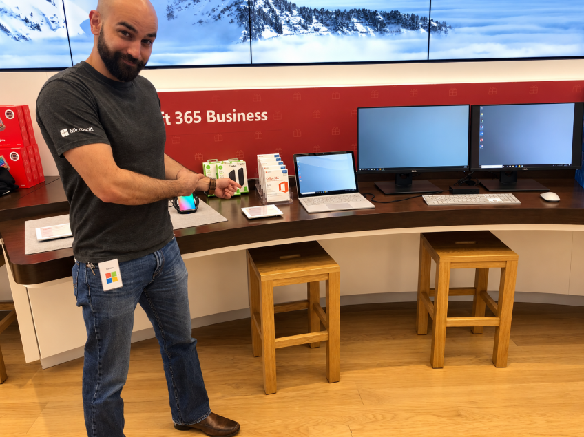 A person gestures toward Microsoft 365 Business products, laptops, and desktop monitors in a showroom.