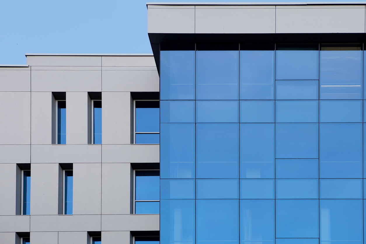 Grey concrete panel facade with narrow vertical windows, blue glass curtain wall, clear blue sky