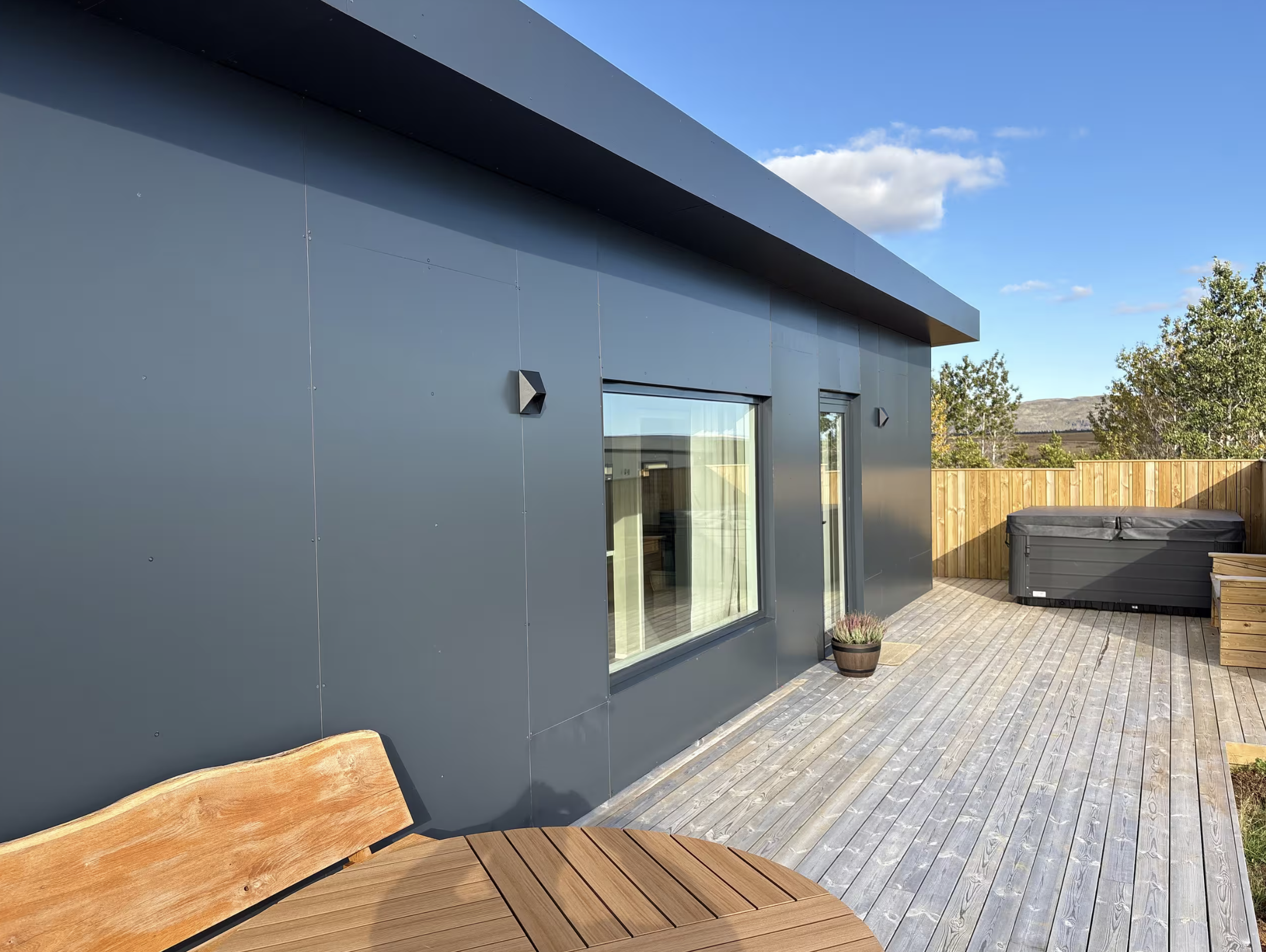 Dark guesthouse exterior with a timber deck, hot tub and trees beyond.